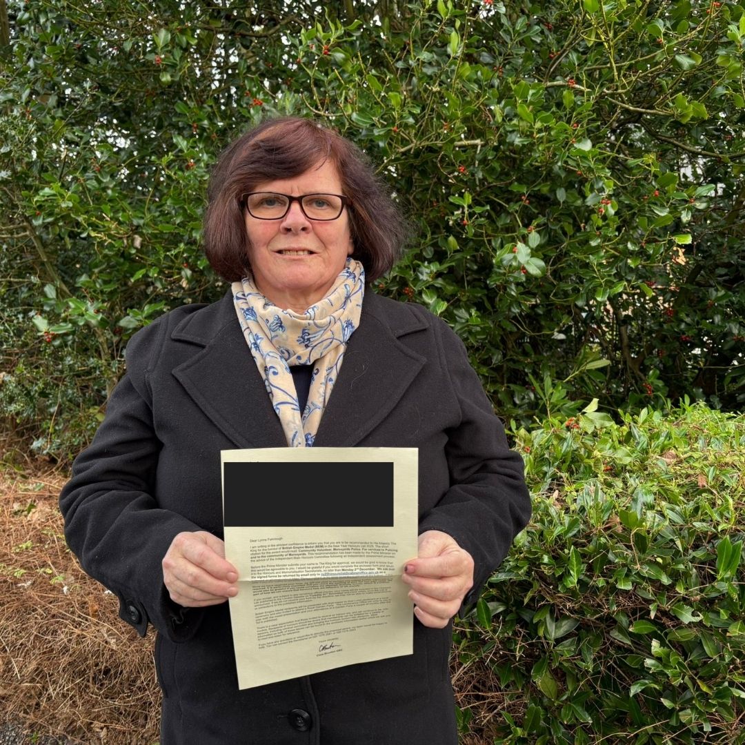 White middle aged woman holds a letter, in front of her garden hedges, smiling at the camera
