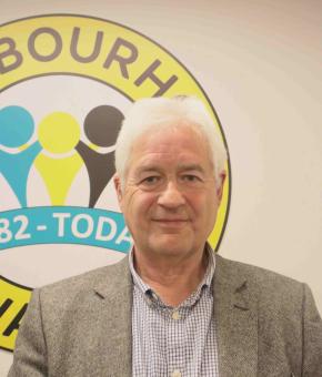 Andrew has white hair, he's wearing a grey jacket and an open neck blue shirt. He's smiling at the camera 